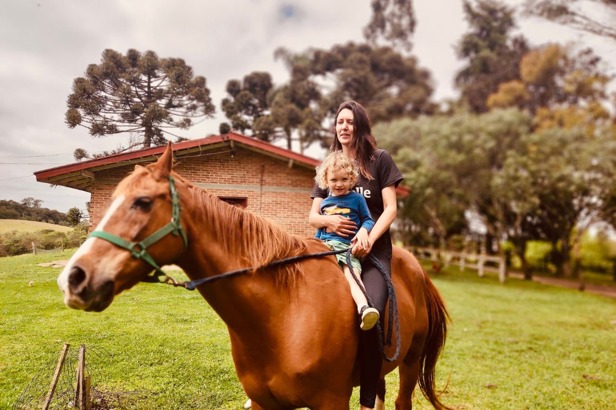 O que é ser mãe atípica? Entenda os desafios que precisam ser superados 2 Heloísa Santos, uma mulher branca de cabelos pretos, com seu filho Oliver, uma criança pequena. Eles estão montados em um cavalo, com uma casa e árvores ao fundo.
